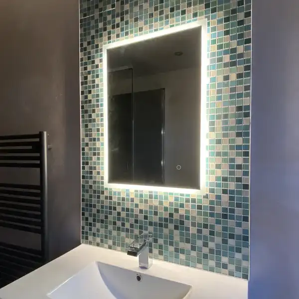 A lit mirror over a white basin, framed by small blue-green tiles