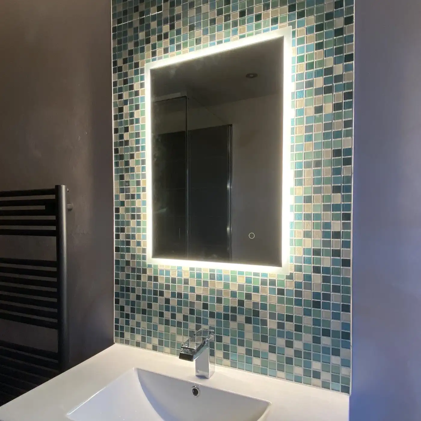 A lit mirror over a white basin, framed by small blue-green tiles