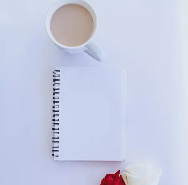 A cup of tea, a blank spiral notebook and two roses on a white table